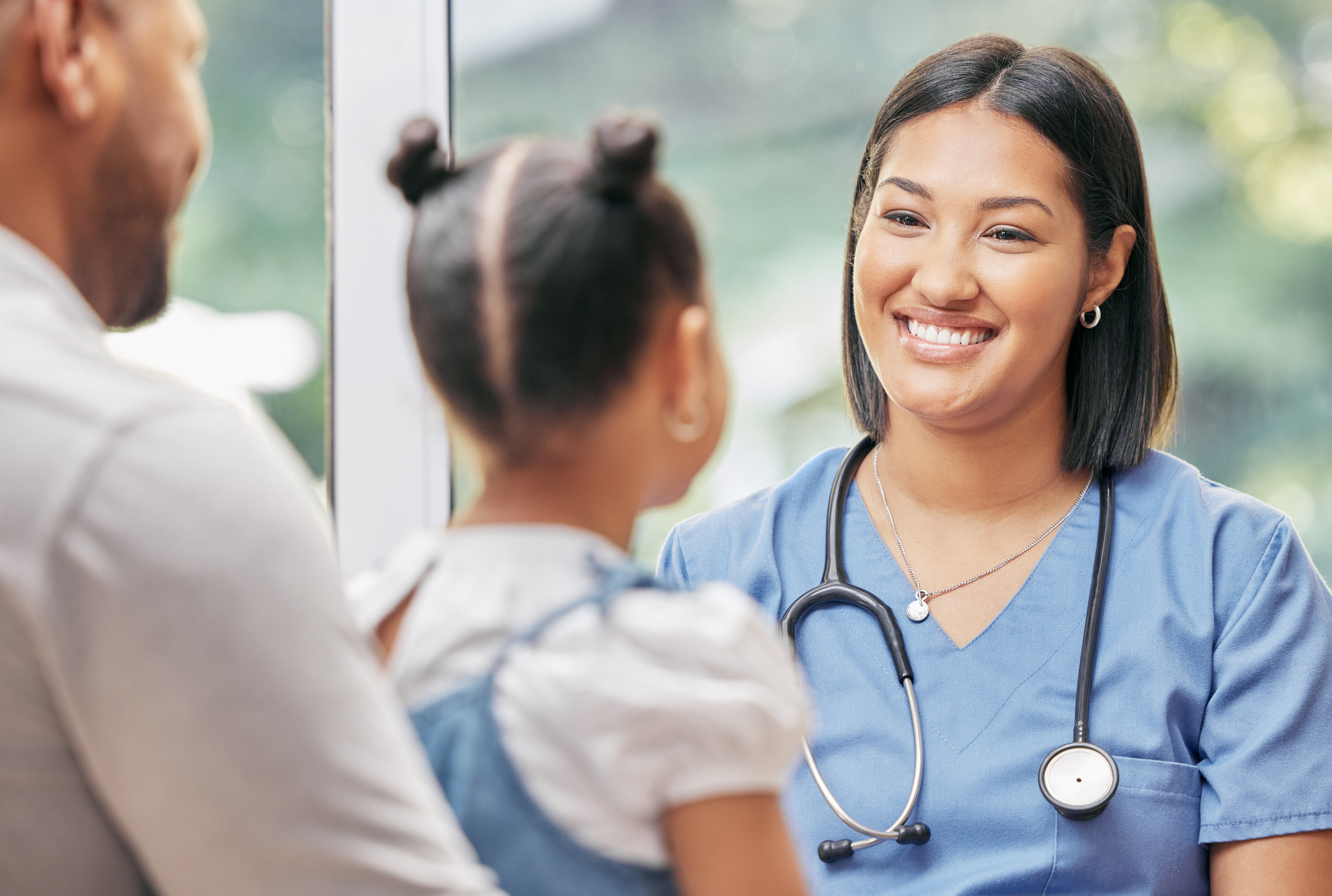 A nurse wearing scrubs with a stethoscope around her neck looks at a young girl who has her hair in buns.