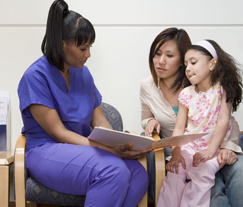 A nurse in blue scrubs speaks with a parent and female child.